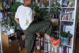 The image shows a person standing in a living space filled with plants and decorative items. They are wearing a loose white top and green pants with white polka dots. One leg is raised, showcasing orange platform sandals. In the background, there is a bookshelf filled with books and various decorative objects, including plants and a poster for "Blade Runner."