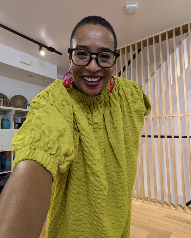 Anita wears a Black Crane turmeric coloured dress with three-quarter length sleeves. She wears magenta Warren Steven Scott earrings