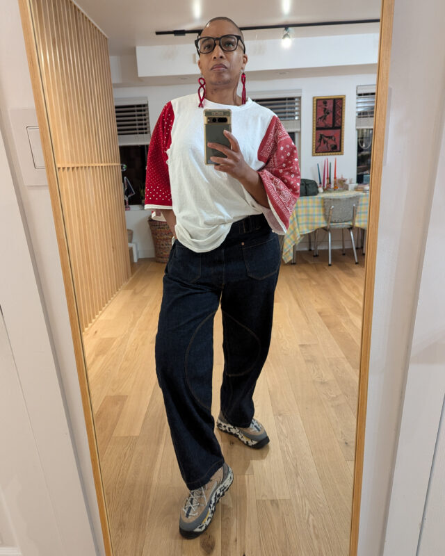 The image shows a person standing in front of a mirror, wearing a loose shirt t-shirt with red raglan sleeves. They're wearing dark indigo Japanese selvage denim. The background features a well-lit room with wooden flooring and a table with a checkered tablecloth.