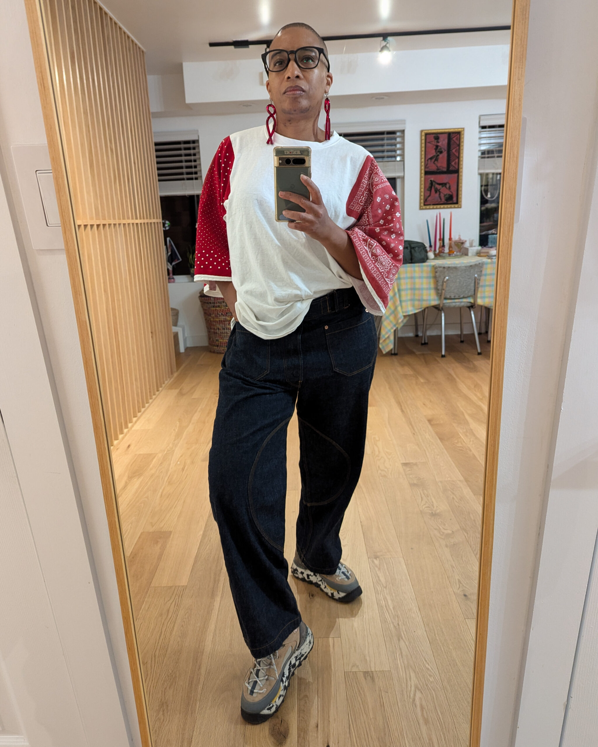 The image shows a person standing in front of a mirror, wearing a loose shirt t-shirt with red raglan sleeves. They have on dark indigo Japanese selvage denim. The background features a well-lit room with wooden flooring and a table with a checkered tablecloth.