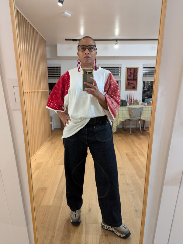 The image shows a person standing in front of a mirror, wearing a loose shirt t-shirt with red raglan sleeves. They're wearing dark indigo Japanese selvage denim. The background features a well-lit room with wooden flooring and a table with a checkered tablecloth.